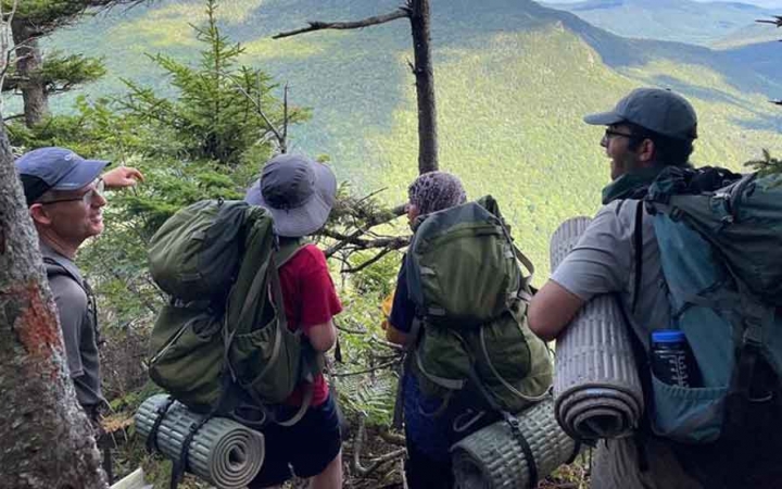 a group of people wearing backpacking gear stand among trees looking out over a green landscape far below